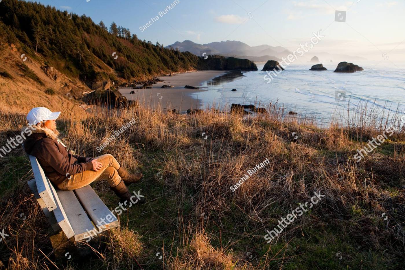 A woman sitting alone on a bench on the headland above Cannon Beach, Oregon, looking out at the ocean at sunset
