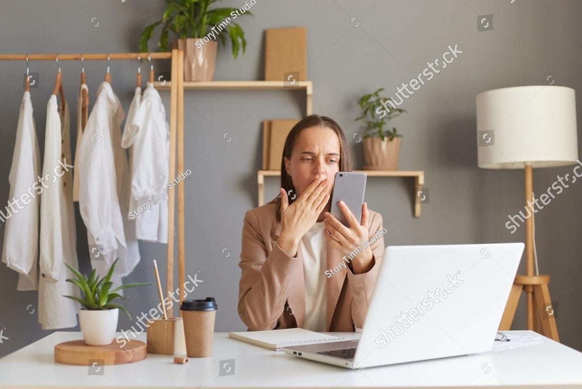 A woman at her desk with her hand over her mouth, looking at her phone